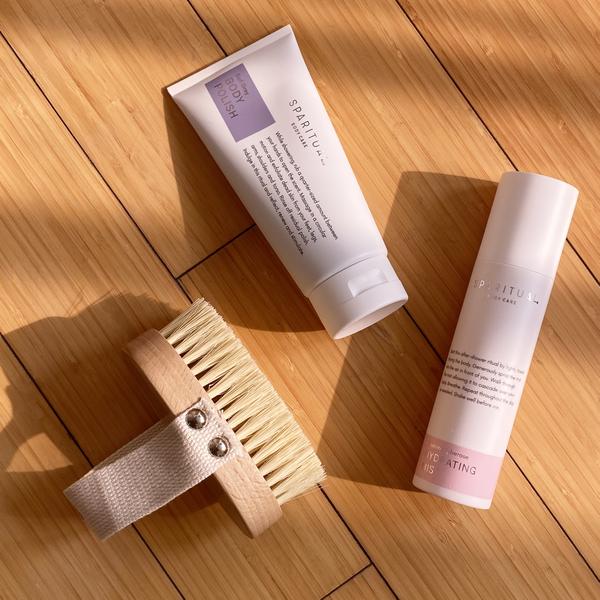 Natural bristle Dry Body Brush | Sparitual with wooden handle and strap, alongside two skincare tubes on a wooden surface in soft sunlight, evoking body care and wellness.
