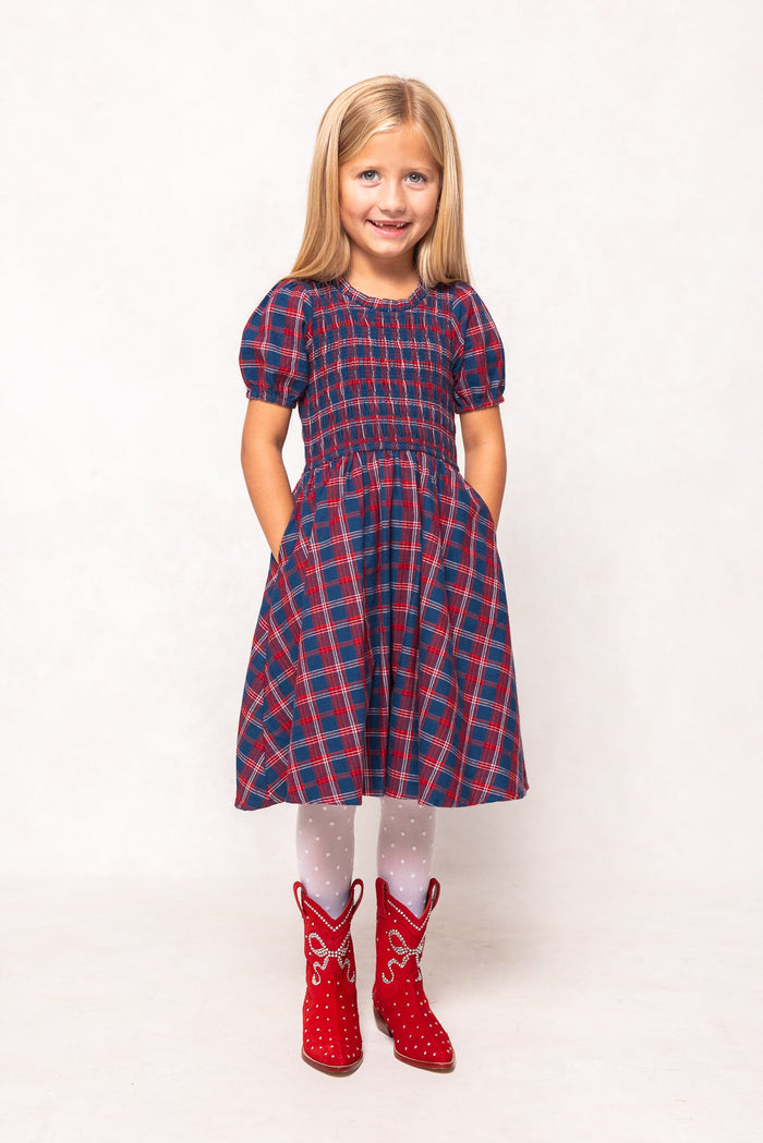 A young girl with long blonde hair smiles as she poses with her hands in her pockets, wearing the Ivy City Co Mini Mariah Dress in Mariner Tartan, paired with white polka-dot tights and red cowboy boots against a plain white background—a charming outfit for any mommy and me occasion.