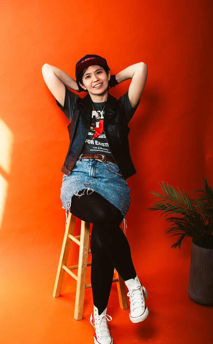 Person sitting on a stool against an orange wall wearing a No Apologies T-shirt from 17th & Granville
