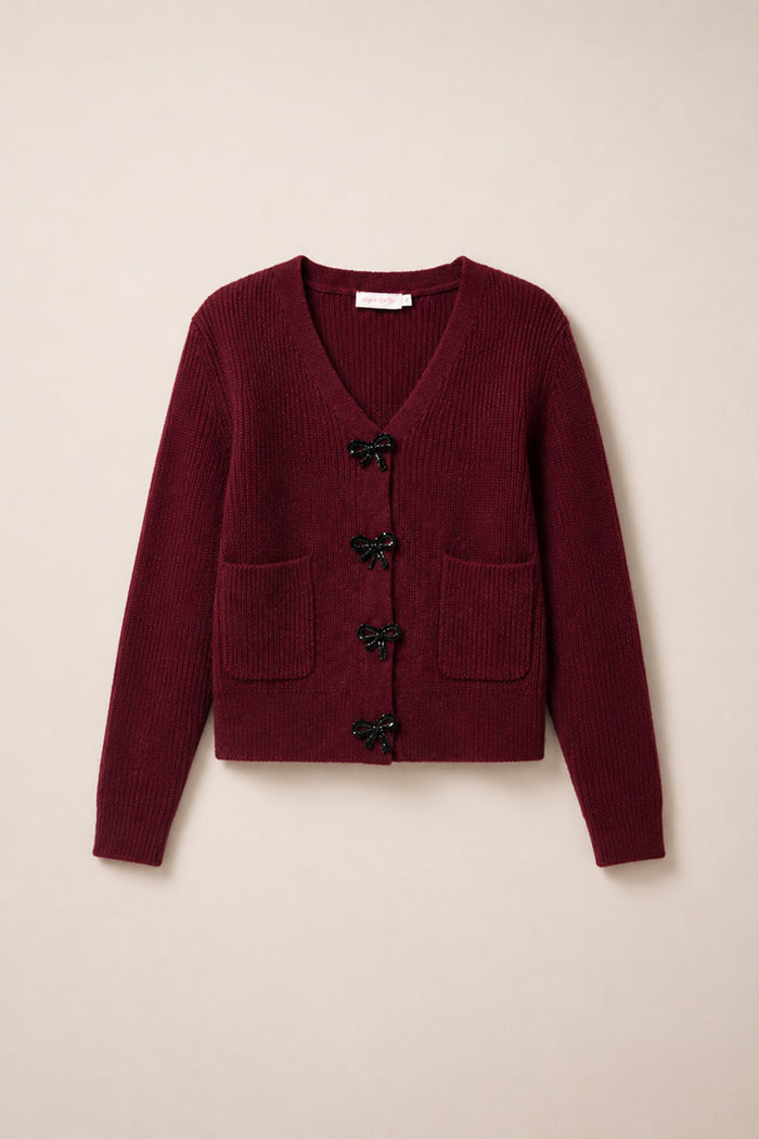The Theo Sweater in Burgundy by Ivy City Co is a cozy fall cardigan featuring a V-neck, two front pockets, and four black bow-shaped button closures down the center. Crafted from an acrylic-nylon blend sweater material, it combines comfort and style—perfect for crisp autumn days.