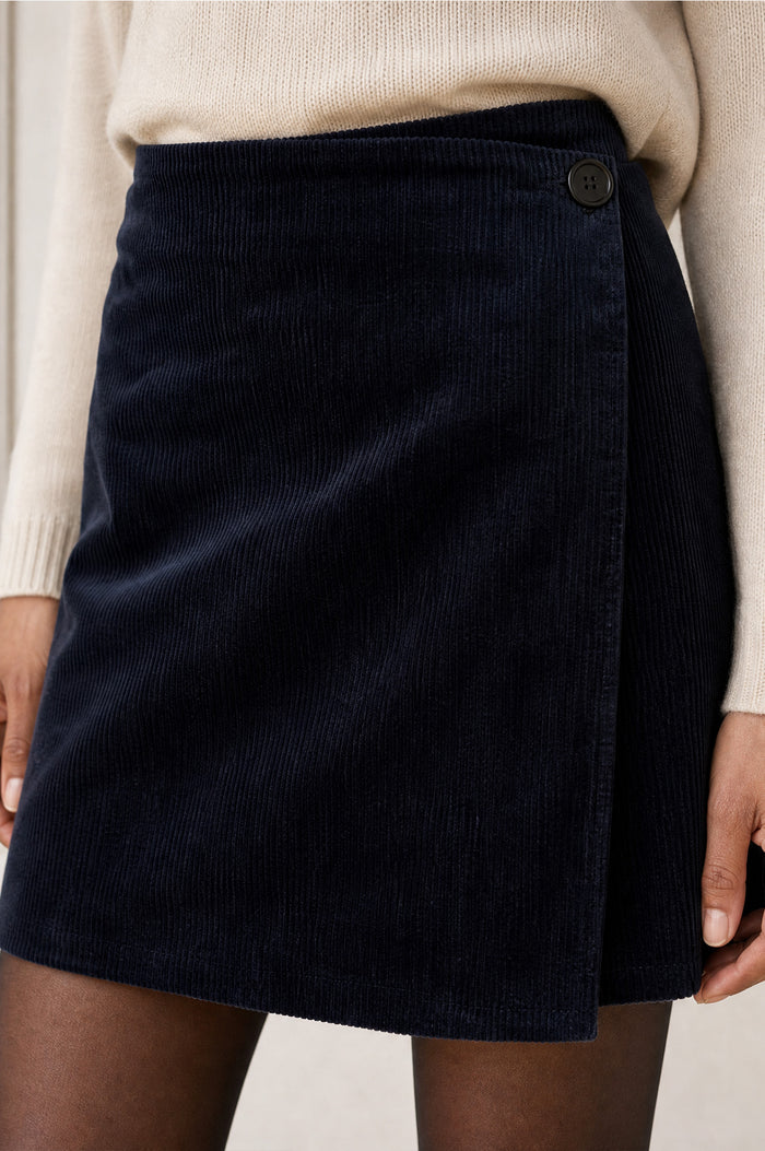 Model wearing the Aspiga Corduroy Wrap Mini Skirt in Dark Navy, a wrap-style corduroy mini skirt with a button fastening, alternate view
