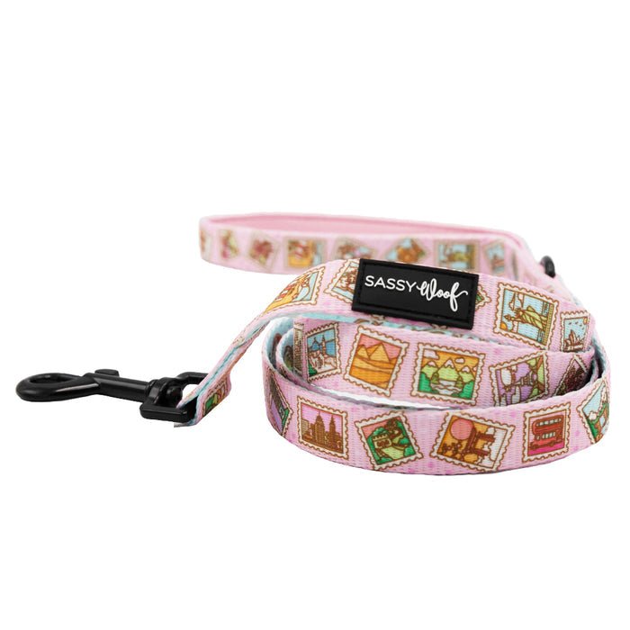 The Sassy Woof Out of Office Dog Leash features pink fabric with colorful stamp graphics, a comfy neoprene handle, sturdy black metal clip, and brand label—perfect for pairing with an adjustable harness for stylish comfort.