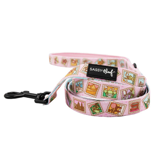 The Sassy Woof Out of Office Dog Leash features pink fabric with colorful stamp graphics, a comfy neoprene handle, sturdy black metal clip, and brand label—perfect for pairing with an adjustable harness for stylish comfort.
