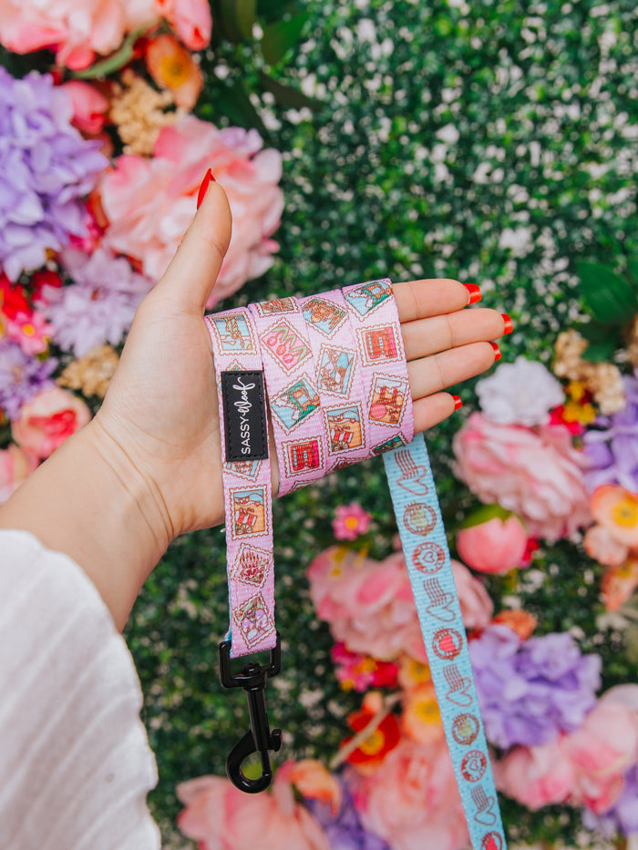 A hand with red nail polish holds the Sassy Woof Dog Leash - Out of Office, featuring playful stamp patterns and a neoprene handle, set against a vibrant backdrop of pink, purple, and white flowers with green foliage.
