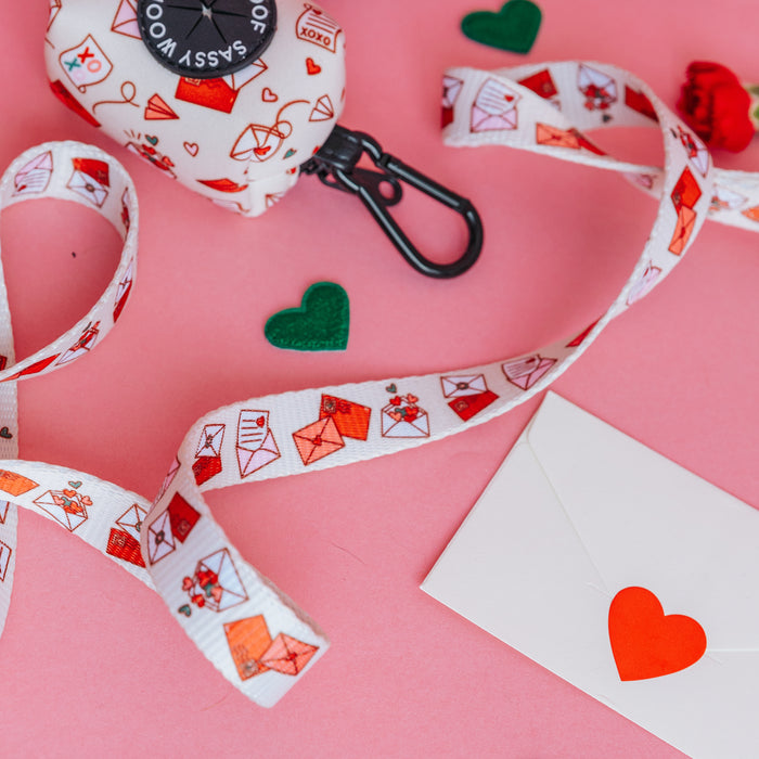 The Sassy Woof Dog Leash - P.S. I Woof You features red and pink envelope designs and is styled on a pink background with heart accents—perfect for pairing with matching harnesses.