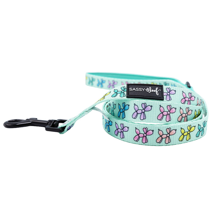The Sassy Woof Pup Pup Hooray dog leash is light blue with colorful balloon dog designs, a black clasp, a branded label, and features a soft neoprene handle for added comfort.