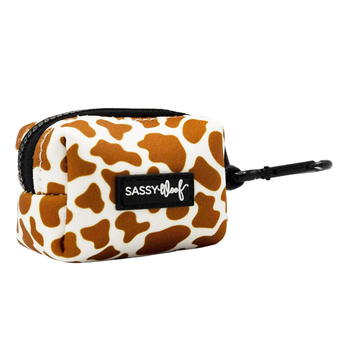 The Sassy Woof Dog Waste Bag Holder - Chocolate Milk features a brown and white cow print, black label, secure zipper, and clip—perfect for adding stylish flair to your cow print dog accessories.