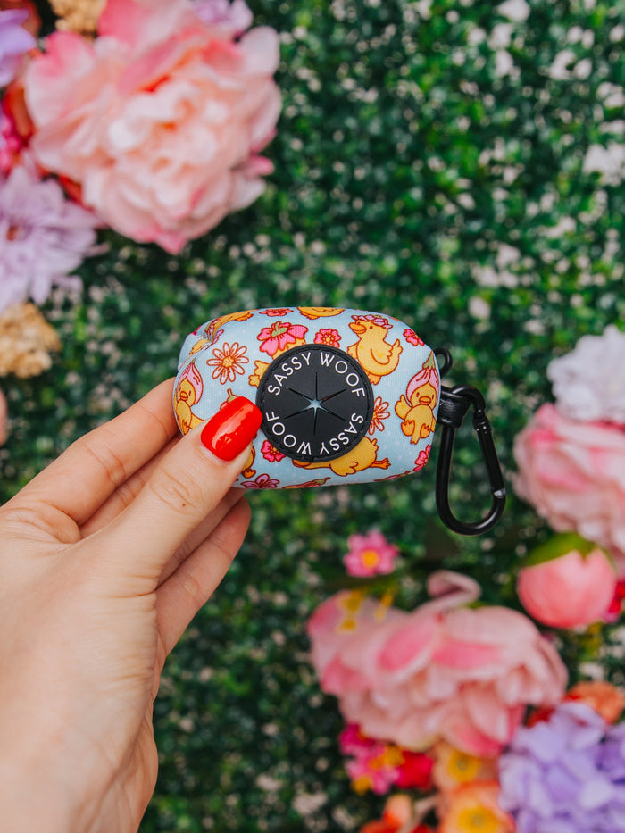 A hand with red-painted nails holds the Sassy Woof "Good Duck, Babe!" neoprene dog waste bag holder featuring a floral and dog design, complemented by lush green foliage and vibrant flowers for a perfect match with any harness.