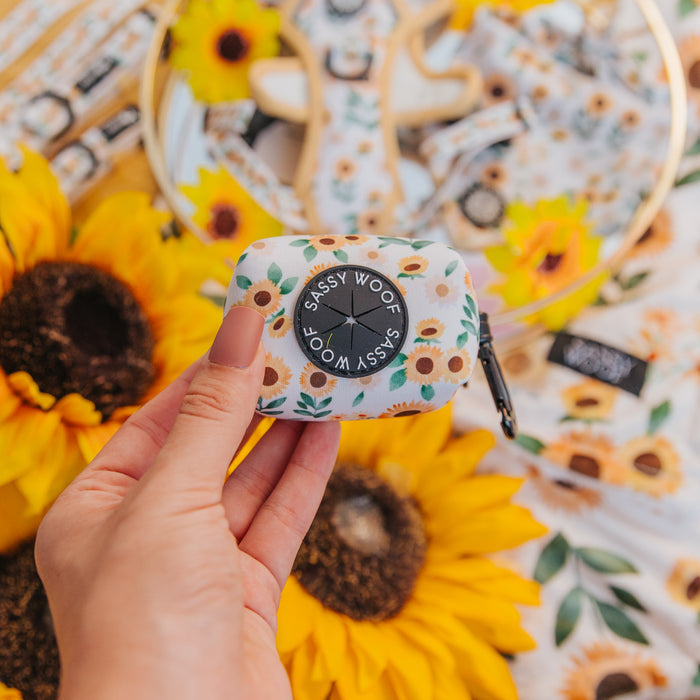 A hand holds the Sassy Woof Dog Waste Bag Holder - Sunflower Dreams, featuring a floral sunflower pattern, with bright yellow sunflowers and a matching harness set in the background.