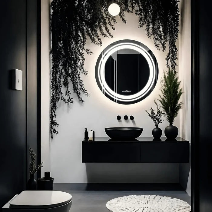 A close-up of a round bathroom LED mirror with a frameless design. The mirror is placed over the grey and black vanity which has a black ceramic sink and some plants with black pots.