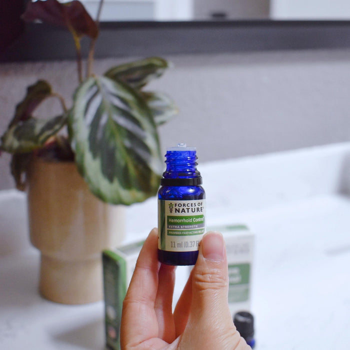 Handheld view of Forces of Nature Hemorrhoid Control 11 ml blue bottle with label, blurred countertop and plant background