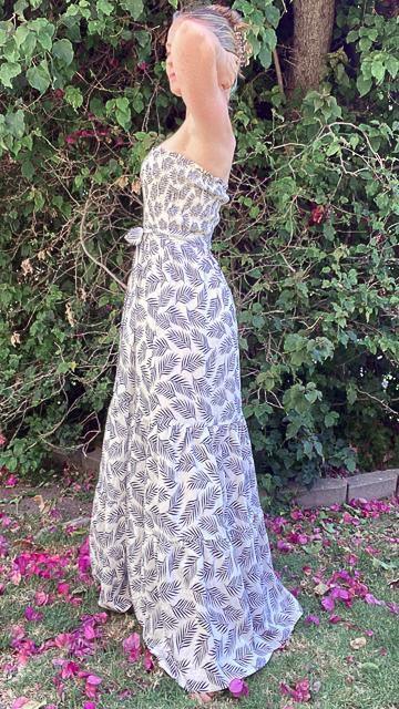 Woman in tropical print smocked maxi dress standing outdoors by leafy green bushes on grass with flower petals