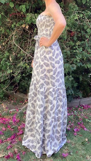 Woman wearing a strapless white maxi dress with black tropical leaf print, standing outdoors with greenery background