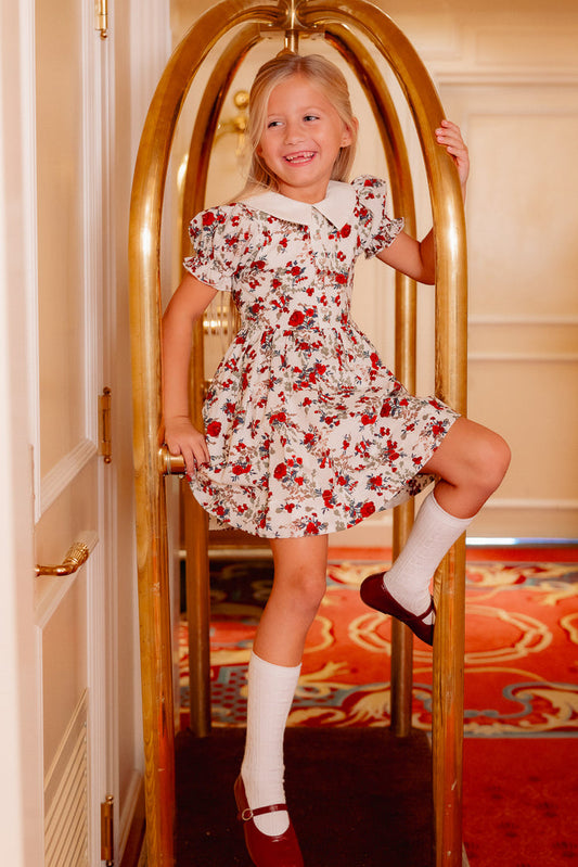 A young girl in the Ivy City Co Mini Aveline Dress in Red & Green Rustic Floral, paired with Mary Jane shoes, smiles as she playfully climbs a gold luggage cart in a hotel hallway with a colorful carpet.