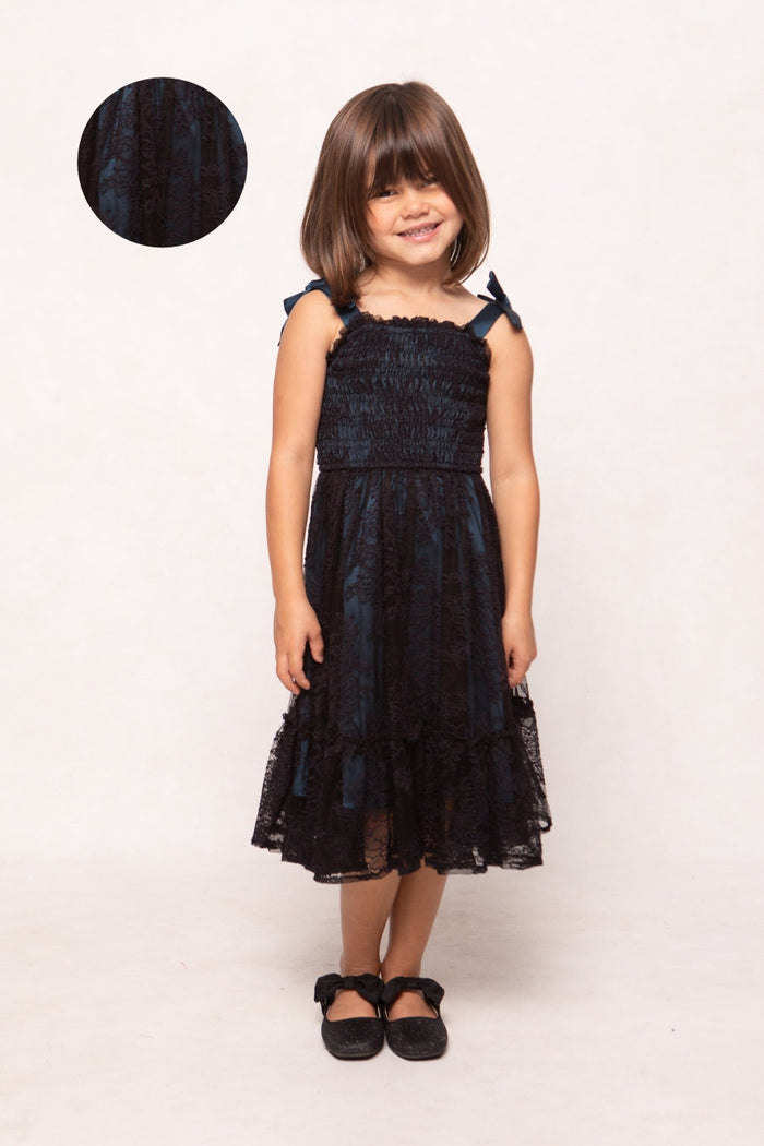 A young girl stands and smiles in the Ivy City Co Mini Isadora Dress in Navy & Black, a sleeveless style featuring bows on the straps. She pairs it with black shoes adorned with bows. An inset displays a close-up of the dress’s fabric pattern, highlighting its suitability for mommy-and-me twinning. Plain white background.