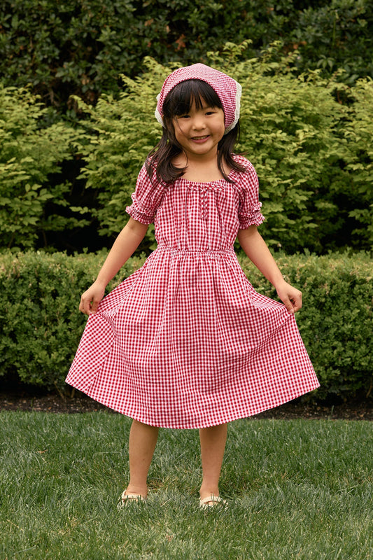 A young girl smiles while holding out her Ivy City Co Mini Jessie Dress in Red Gingham, paired with a matching headscarf, standing on grass with green bushes in the background—perfect for a festive Fourth of July look.