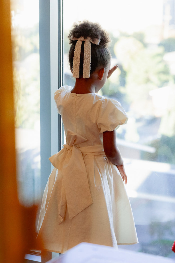 A young girl with curly hair tied in a white ribbon, wearing the Ivy City Co Mini Wrenley Dress in Cream Taffeta, stands with her back to the camera by a window, gazing out at a sunlit outdoor scene.