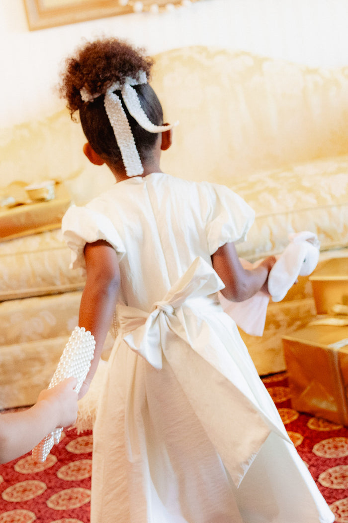 A young girl wearing the Ivy City Co Mini Wrenley Dress in Cream Taffeta with a large bow walks on a red patterned carpet, holding accessories and surrounded by gold-wrapped presents. Her hair is styled with beaded ribbons.