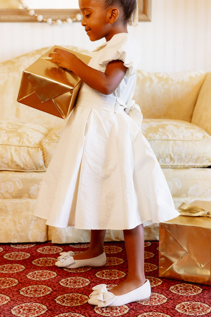 A young girl in the Ivy City Co Mini Wrenley Dress in Cream Taffeta and white shoes smiles on a red carpet, holding a gold-wrapped gift, with another gold present nearby and a cream-colored sofa in the background.