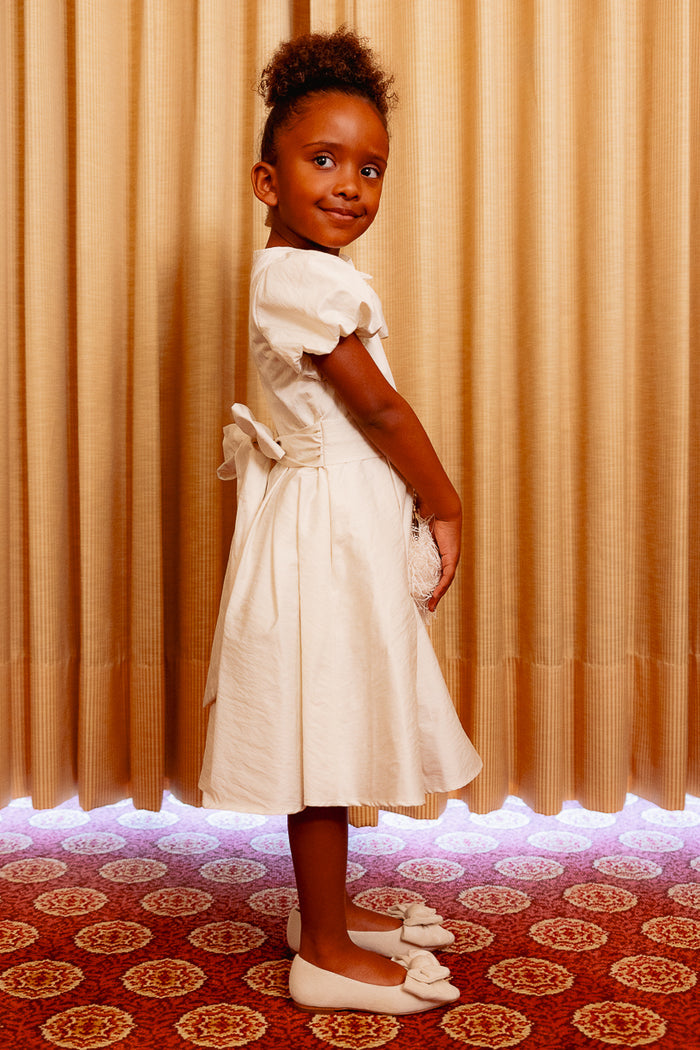 Little girl wearing a white dress standing sideways against a yellow curtain and on a red carpet