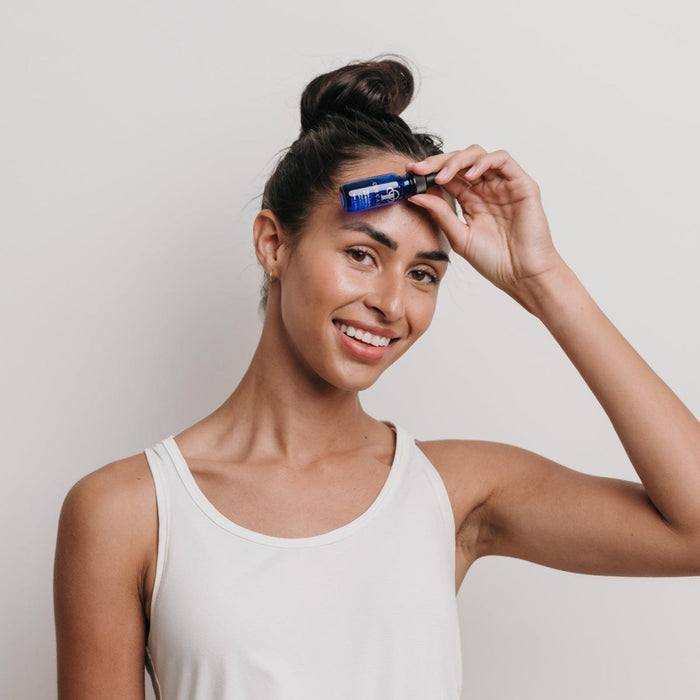Young woman in a white tank top smiling and holding a small blue bottle of Myo-Cyte Plus | Circadia skincare serum near her forehead in a wellness skincare setting.