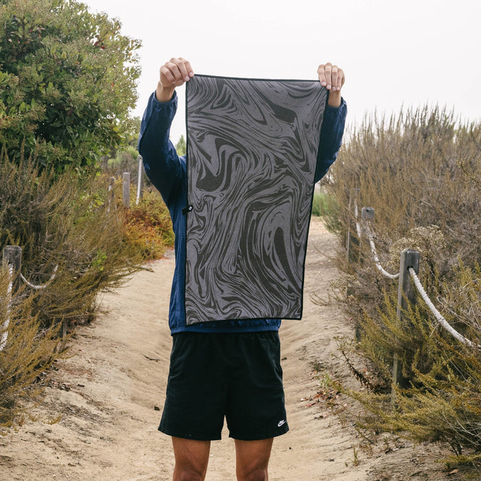 Person holding a grey and black marbled Nomadix fitness towel on a sandy outdoor trail.