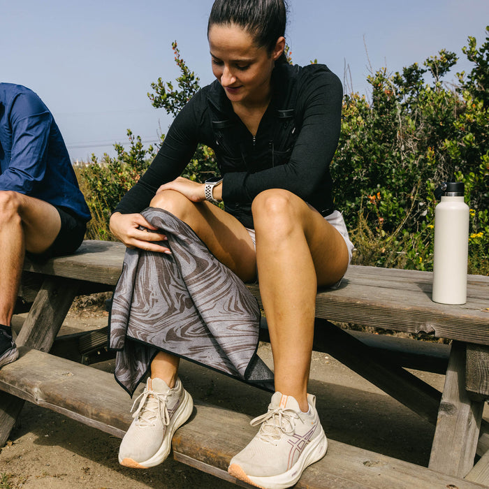 Woman on a wooden bench holding a grey and black marbled Nomadix fitness towel made from recycled materials.