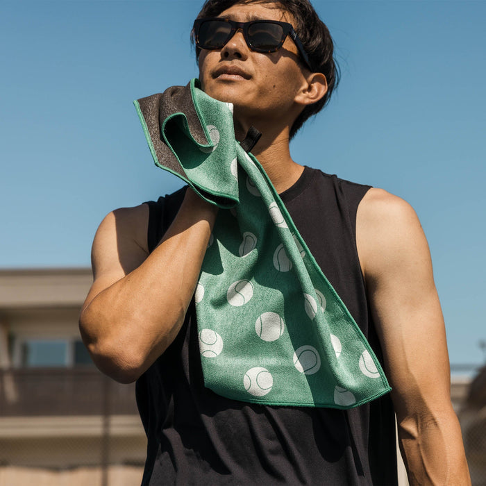 Man wiping sweat with a green, tennis-ball-patterned Nomadix fitness towel made from recycled materials.