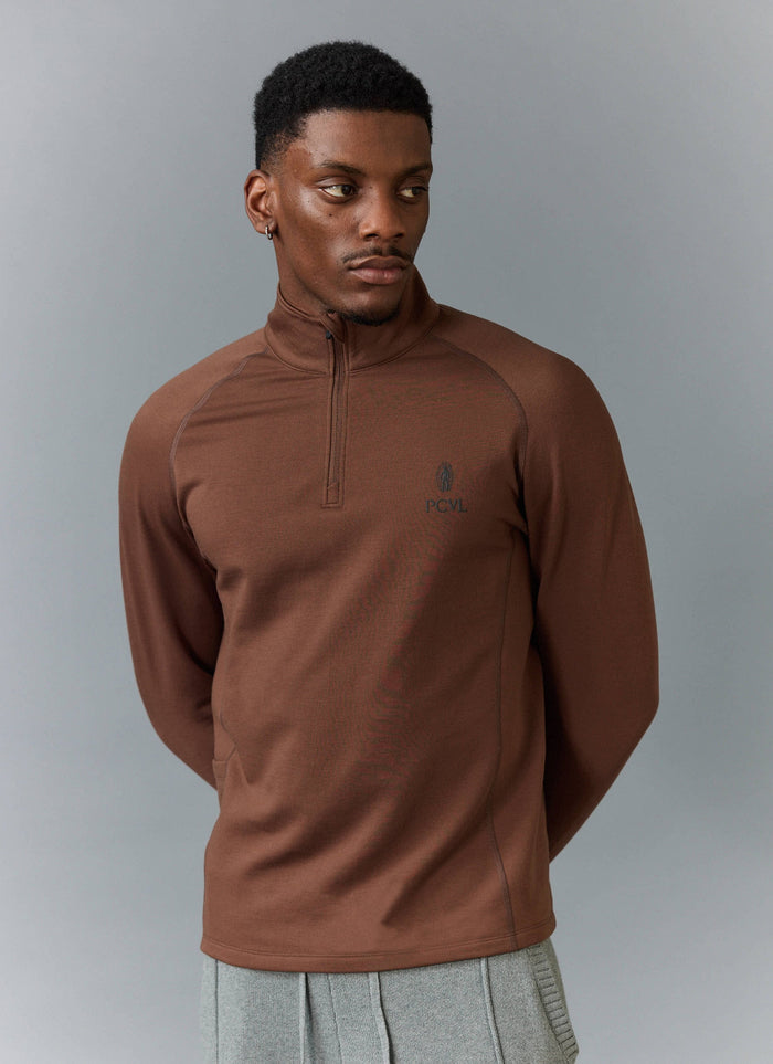 A model wears the Percival PCVL Base Layer Quarter Zip Jacket in rust, showing the zip funnel neck and chest logo.