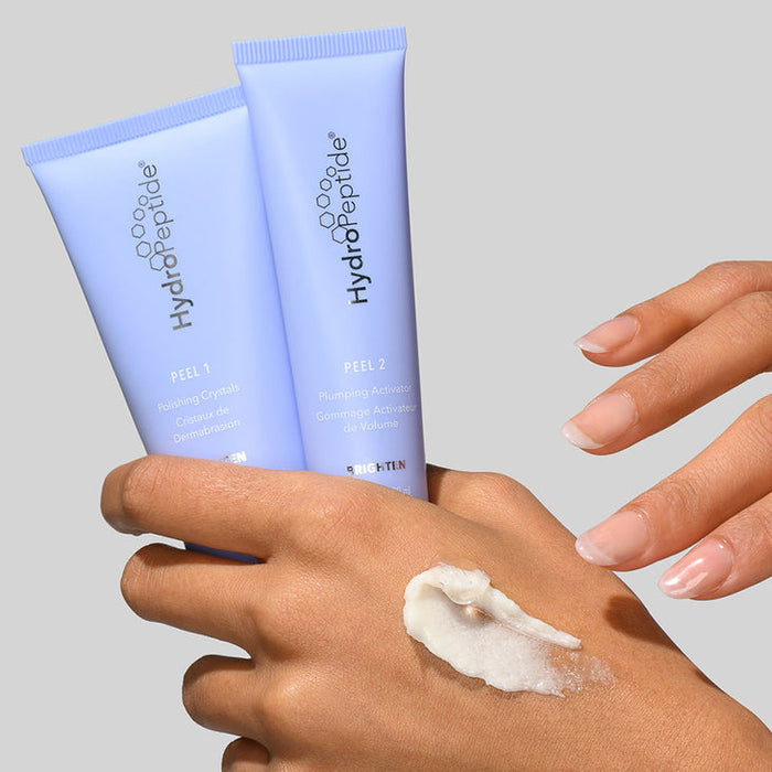 Close-up of hands holding two light purple tubes of Polish & Plump Face Peel Kit | HydroPeptide skincare product with a small amount of white peel cream applied on the back of one hand.
