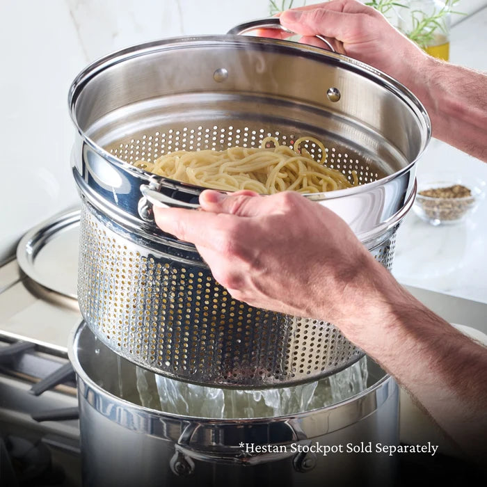 Person holding a metal colander with pasta over a pot on a stove.