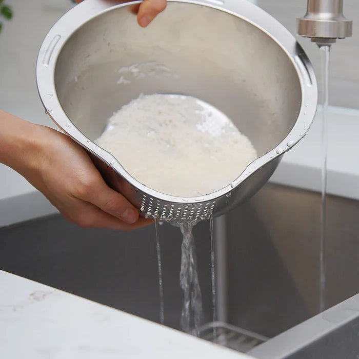 Person rinsing rice in a metal colander under running water in a kitchen sink.