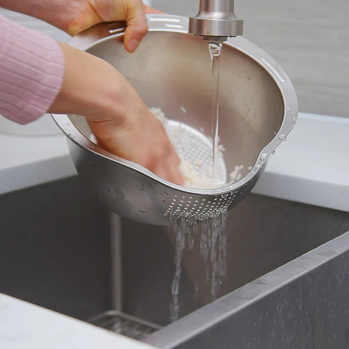 Person washing rice in a metal colander under running water in a kitchen sink.