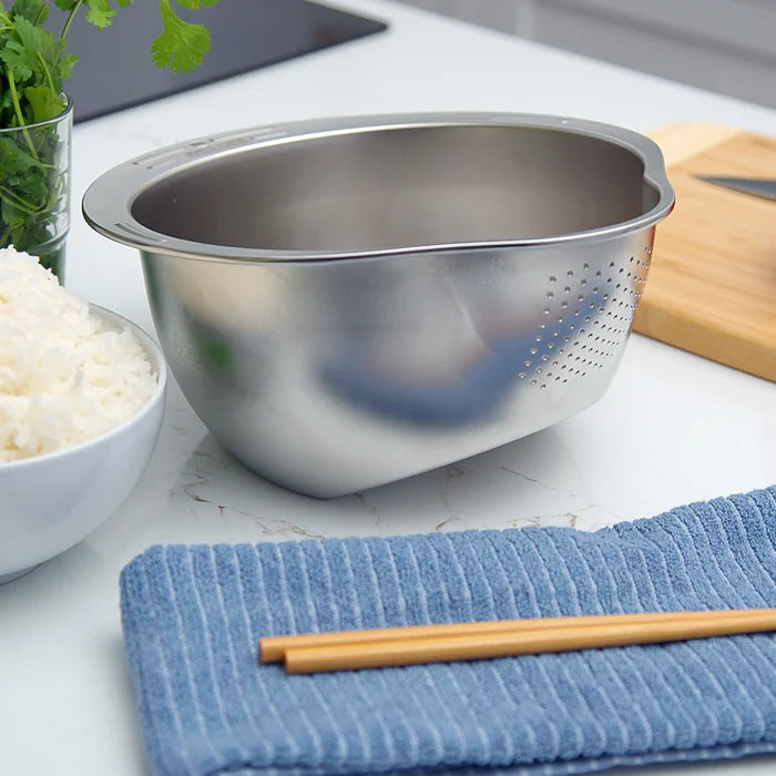 Stainless steel colander on a kitchen counter with rice, a towel, and chopsticks.