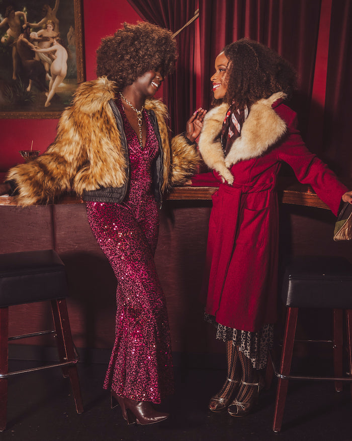 Two women in glamorous outfits - a studio 54 style disco jumpsuit and a fur bombet jacket standing in a bar with red walls and artwork.