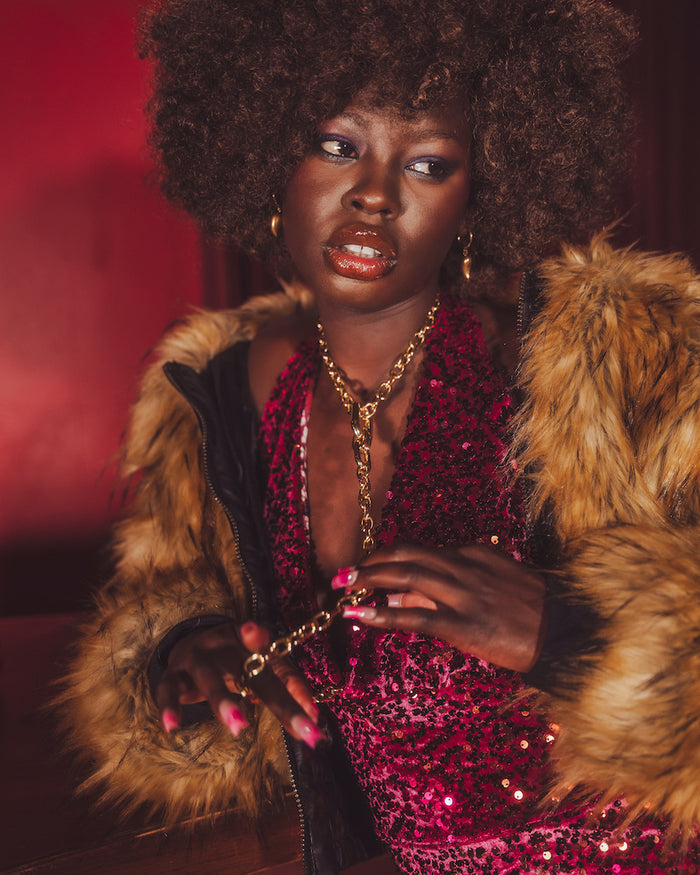 Disco Babe in a retro inspired 70's fur coat and sequin jumpsuit holding a chain, against a red background