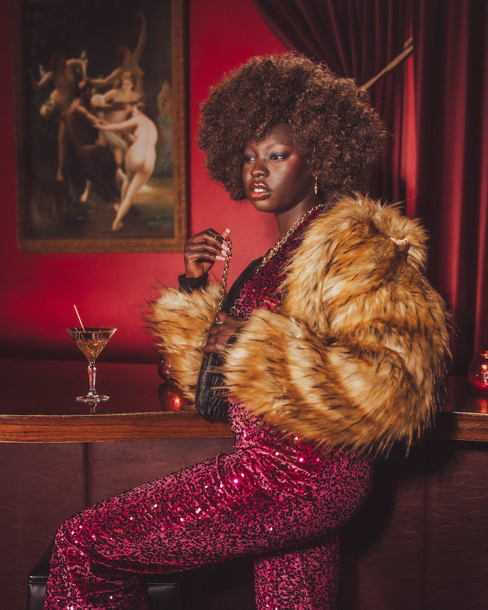 Woman in a glamorous setting with a 1970s style fur coat and cocktail, against a red wall with a painting.