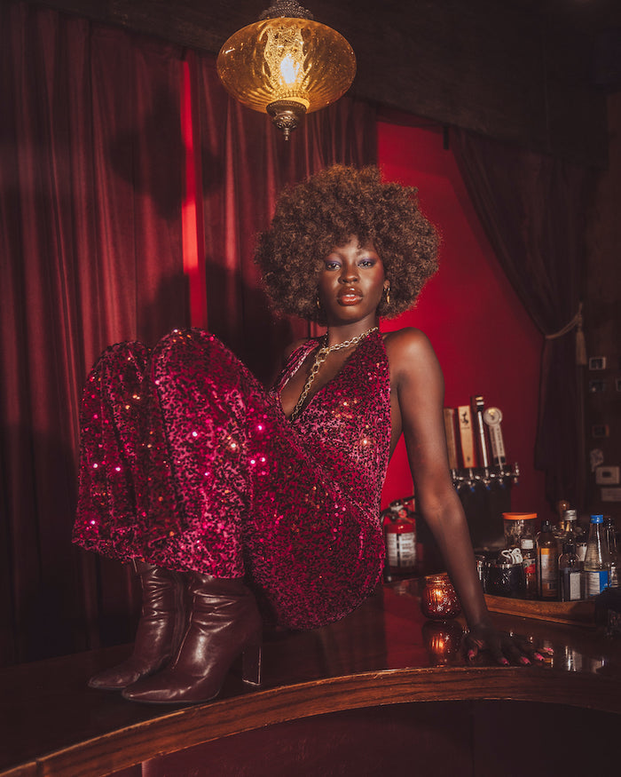 Woman in a red sequin jumpsuit in a 70s retro bar standing behind a table with red curtains and lighting.
