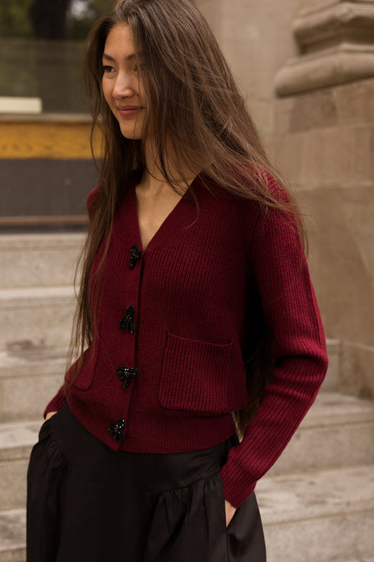 A woman wearing the Theo Sweater in Burgundy by Ivy City Co—a cozy acrylic-nylon knit cardigan with black bow embellishments and front pockets—paired with a black skirt, stands on stone steps outdoors.