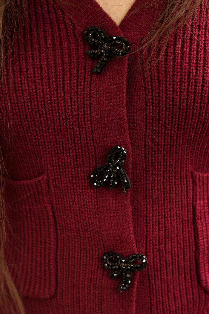 Close-up of the Theo Sweater in Burgundy by Ivy City Co, a cozy acrylic-nylon blend in a rich maroon knit. The sweater features three black beaded bow buttons and two small front pockets, while strands of long brown hair along the sides add a soft touch.