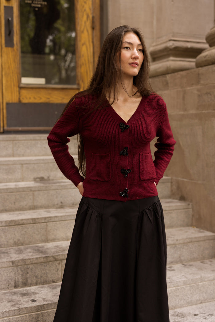 A woman with long brown hair stands on stone steps, wearing the Theo Sweater in Burgundy by Ivy City Co paired with a long black skirt. A wooden door and stone building create an inviting backdrop.