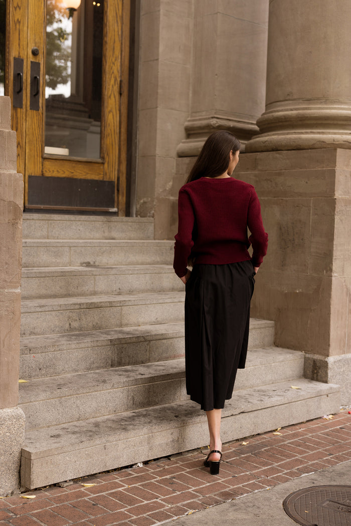 Wearing the Theo Sweater in Burgundy by Ivy City Co, a woman with her hands in her pockets walks up stone steps toward large wooden doors, flanked by tall columns, pairing her cozy fall knit with a black skirt.