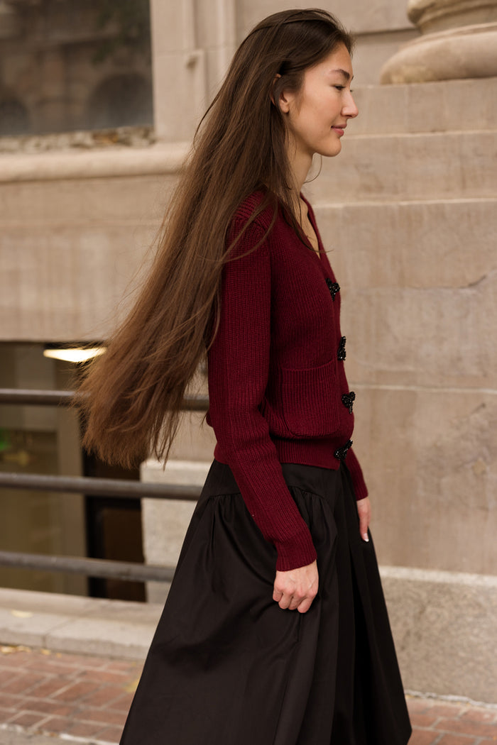 A woman with long brown hair, dressed in Ivy City Co’s Theo Sweater in Burgundy and a black skirt, walks outdoors beside a stone building.