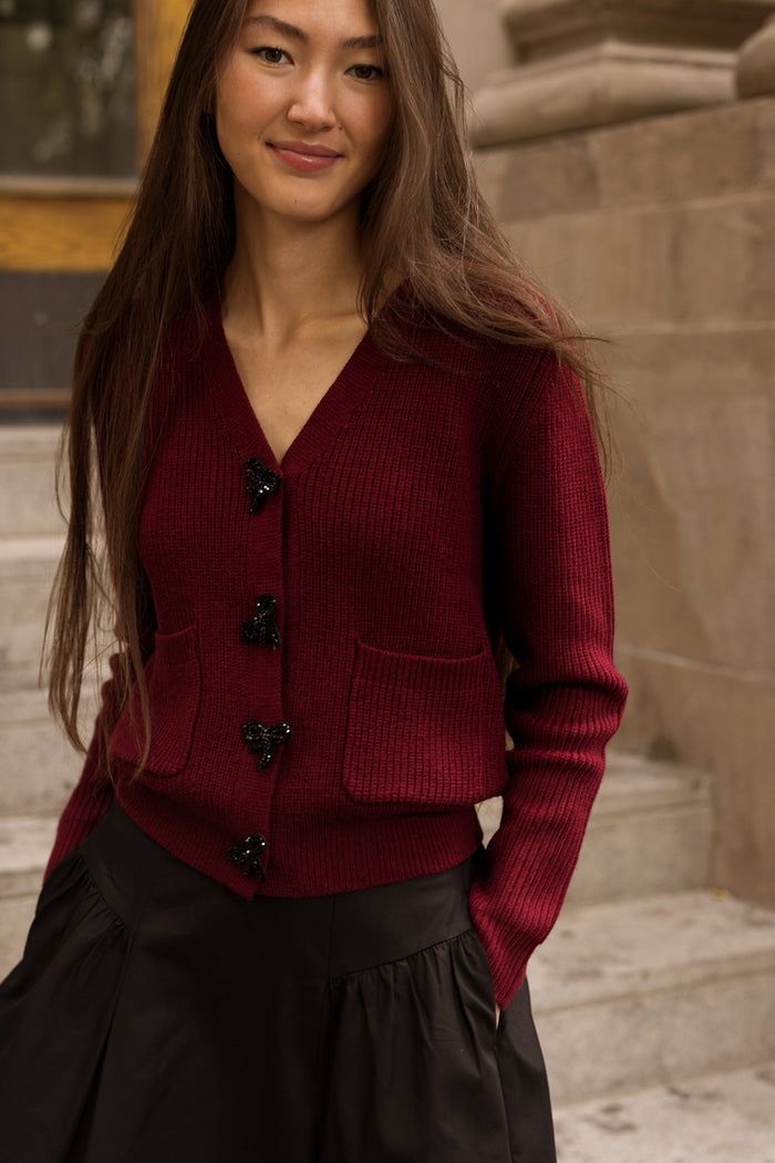 A woman stands outdoors on stone steps, smiling softly at the camera. She wears Ivy City Co’s Theo Sweater in Burgundy—a cozy fall cardigan with black buttons and pockets—paired with a black skirt. Her long brown hair frames her face as she enjoys the crisp autumn day.