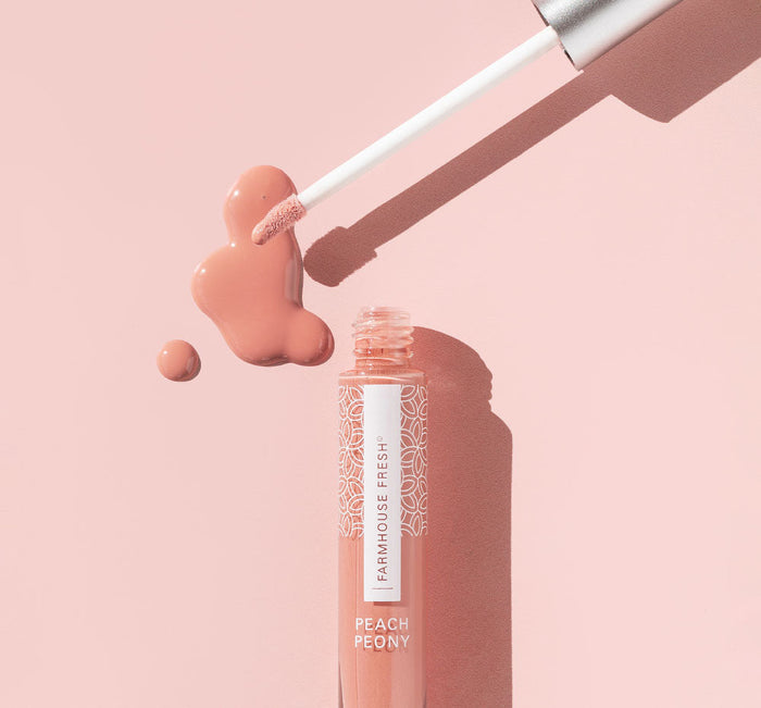 An open tube of Vitamin Glaze® Oil Infused Lip Gloss – Peach Peony | Farmhouse Fresh with a wand applicator hovering above a glossy peach-colored spill on a soft pink background.