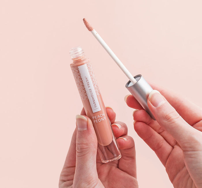 Hands holding a tube and applicator of Vitamin Glaze® Oil Infused Lip Gloss – Peach Peony | Farmhouse Fresh with peachy nude gloss on a soft pink background