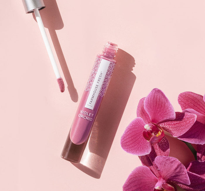 Vitamin Glaze® Oil Infused Lip Gloss – Violet Orchid | Farmhouse Fresh with a shimmery mauve shade shown alongside violet orchid flowers on a soft pink background in a skincare and beauty setting.
