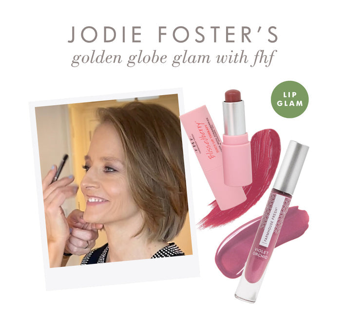 Jodie Foster smiling while applying makeup, alongside a display of Vitamin Glaze® Oil Infused Lip Gloss – Violet Orchid | Farmhouse Fresh with pink and violet lip colors shown.